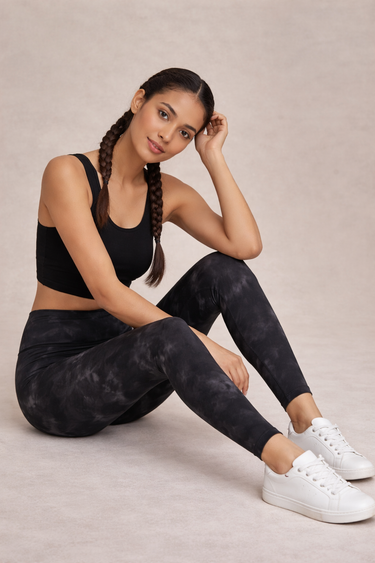 Woman wearing black tie-dye leggings and a black sports bra, sitting on a light gray surface.