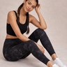 Woman wearing black tie-dye leggings and a black sports bra, sitting on a light gray surface.