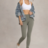 Woman wearing a white sports bra and khaki green leggings with a denim jacket on a plain background