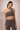 Woman wearing a brown one-shoulder scallop detail sports bra and scallop detail leggings against a beige background